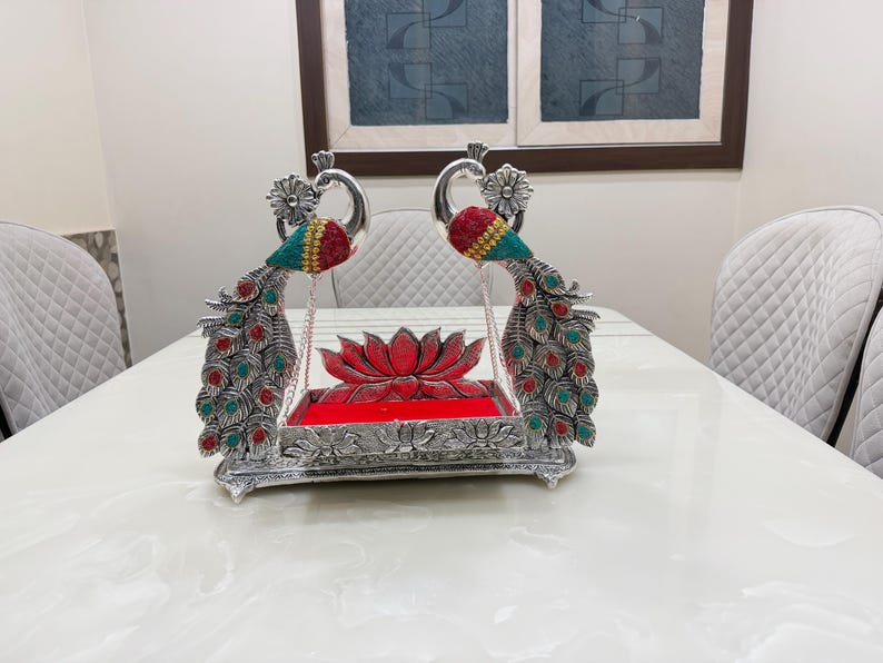 May include: A silver-toned decorative swing featuring two peacock figures with red and teal accents. The base is rectangular with a lotus flower motif, and the central platform is lined with red fabric, suspended by thin silver chains.