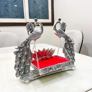May include: A silver-toned decorative swing featuring two peacock figures with detailed plumage. The swing is suspended by metal chains from the peacocks and features a rectangular base with a bright red velvet cushion and a lotus flower accent.