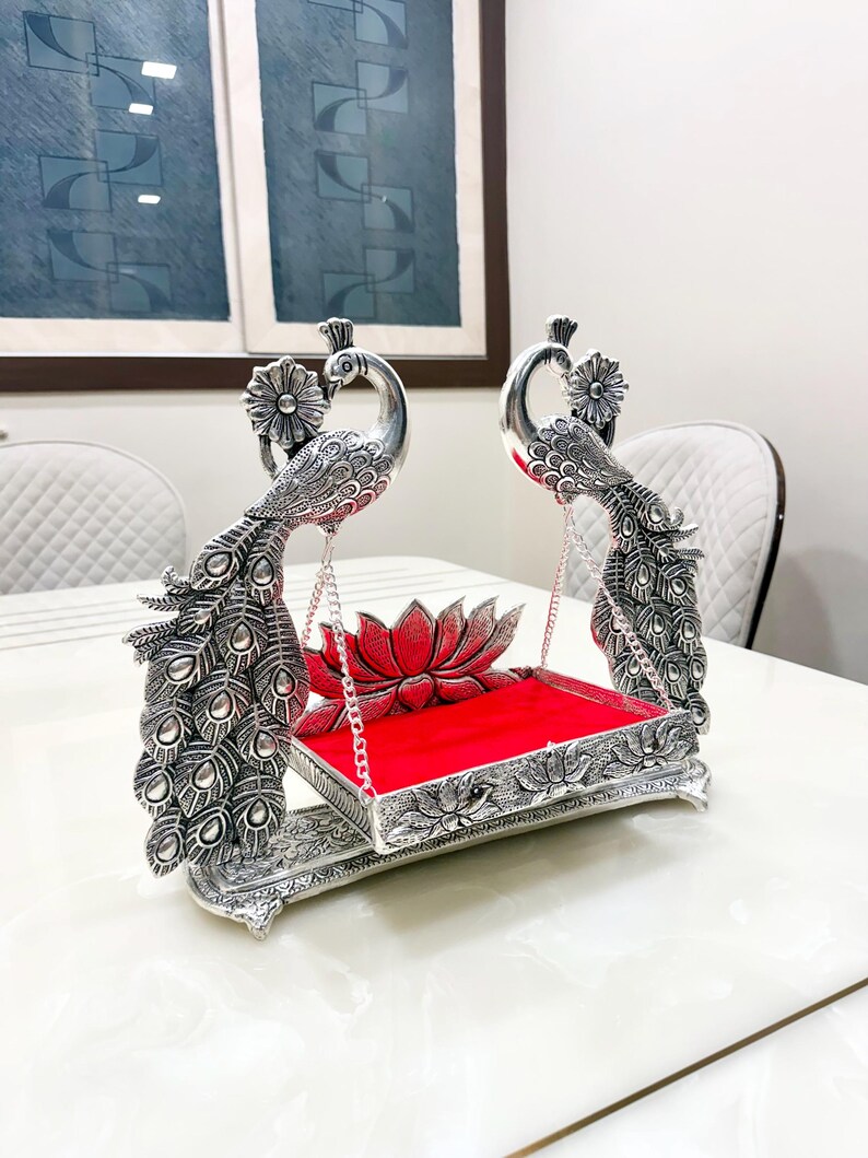 May include: A silver-toned decorative swing featuring two peacock figures with detailed plumage. The swing is suspended by metal chains from the peacocks and features a rectangular base with a bright red velvet cushion and a lotus flower accent.