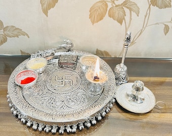 Peut inclure: Un thali de puja en argent avec un motif de swastika, un petit brûleur d'encens, un diya avec une bougie allumée et une petite boîte avec un couvercle. Le thali est décoré de motifs floraux complexes.