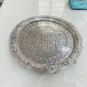 May include: A silver tray with intricate floral designs and a raised swastika symbol in the center. The tray has a scalloped edge and a beaded border.