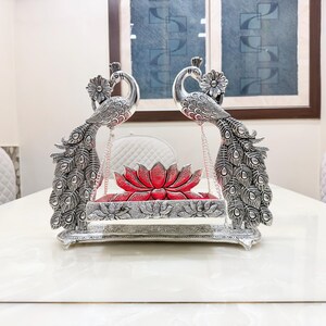 May include: A silver-toned metal swing featuring two ornate peacock figures facing each other, with detailed feather patterns and floral accents. A red lotus-shaped cushion sits on the central platform, which is suspended by thin metal chains.