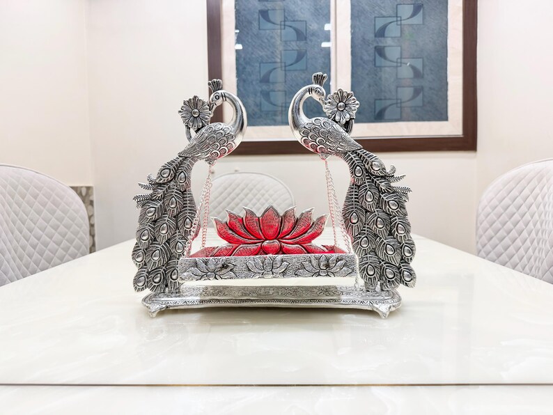 May include: A silver-toned metal swing featuring two ornate peacock figures facing each other, with detailed feather patterns and floral accents. A red lotus-shaped cushion sits on the central platform, which is suspended by thin metal chains.