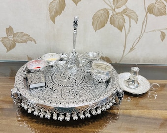 May include: A silver-plated puja thali with intricate floral designs and a swastika symbol in the center. The thali includes a bell, a diya, a small incense burner, and other puja items.