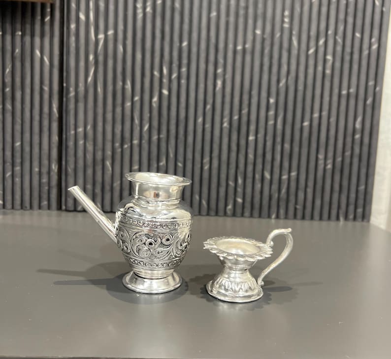 May include: Two silver decorative containers with intricate floral designs. The larger container has a spout and a round base. The smaller container has a handle and a round base.
