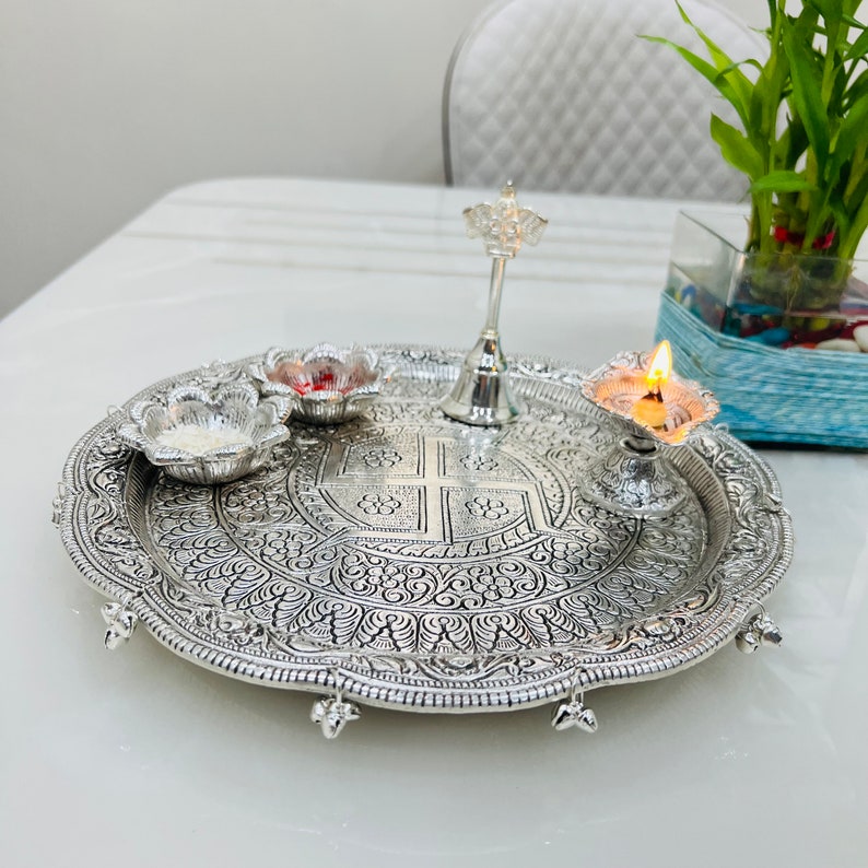 May include: A silver tray with intricate designs, featuring a small silver bell, a diya with a lit candle, and two small bowls. The tray has a scalloped edge and decorative feet.