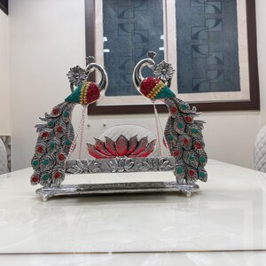 May include: A silver-toned decorative swing featuring two peacock figures with red and teal accents. The peacocks flank a central red lotus flower ornament suspended by thin chains above a rectangular base with embossed floral patterns.