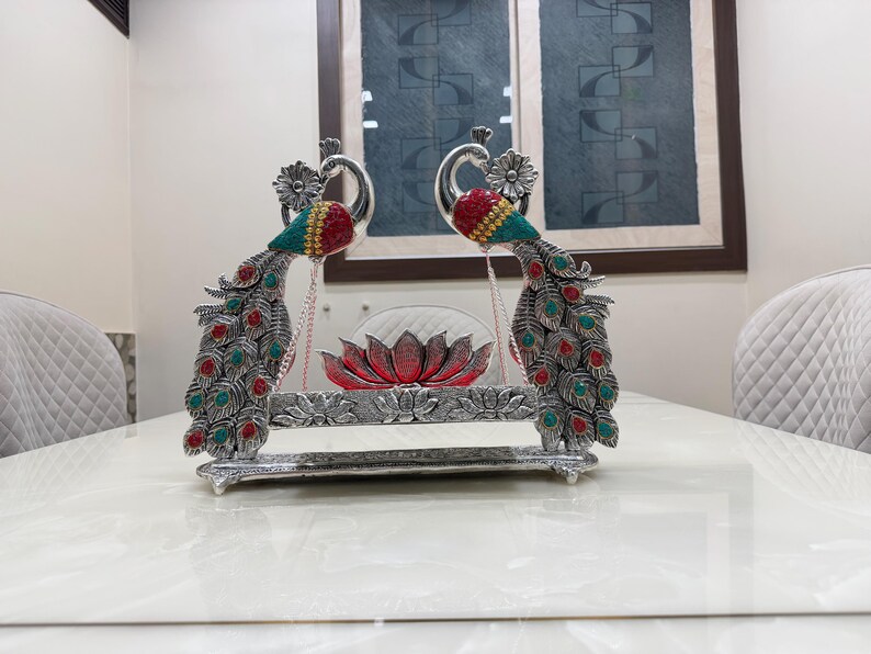 May include: A silver-toned decorative swing featuring two peacock figures with red and teal accents. The peacocks flank a central red lotus flower ornament suspended by thin chains above a rectangular base with embossed floral patterns.