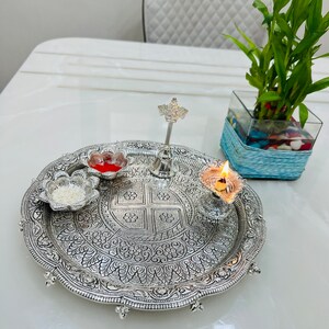 May include: A silver tray with intricate floral designs, featuring a small silver bell, a red and white flower-shaped dish, a white flower-shaped dish with white rice, and a silver oil lamp with a lit wick.