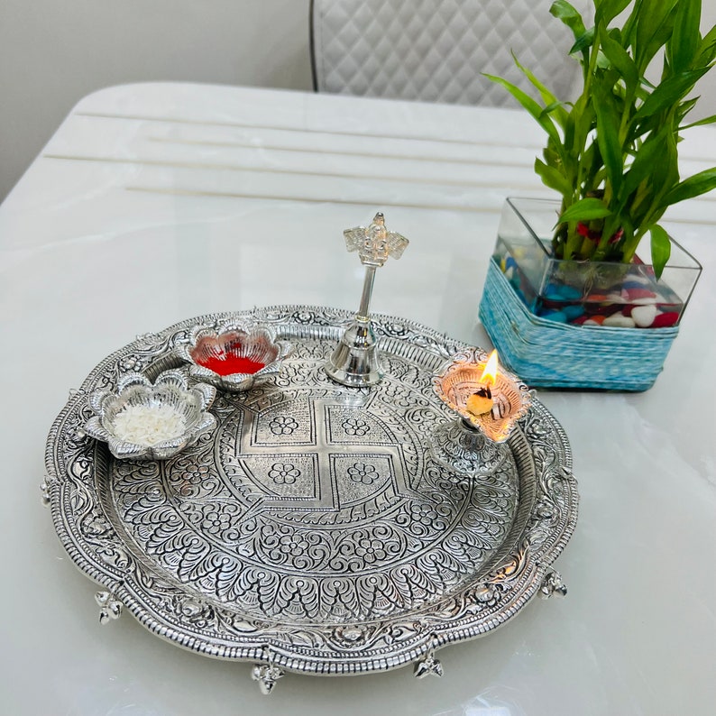 May include: A silver tray with intricate floral designs, featuring a small silver bell, a red and white flower-shaped dish, a white flower-shaped dish with white rice, and a silver oil lamp with a lit wick.