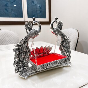 May include: A silver-colored metal swing featuring two peacock figures with detailed feather patterns on the sides. The swing has a rectangular base with a bright red velvet-like insert and a lotus flower motif at the back, suspended by chains.