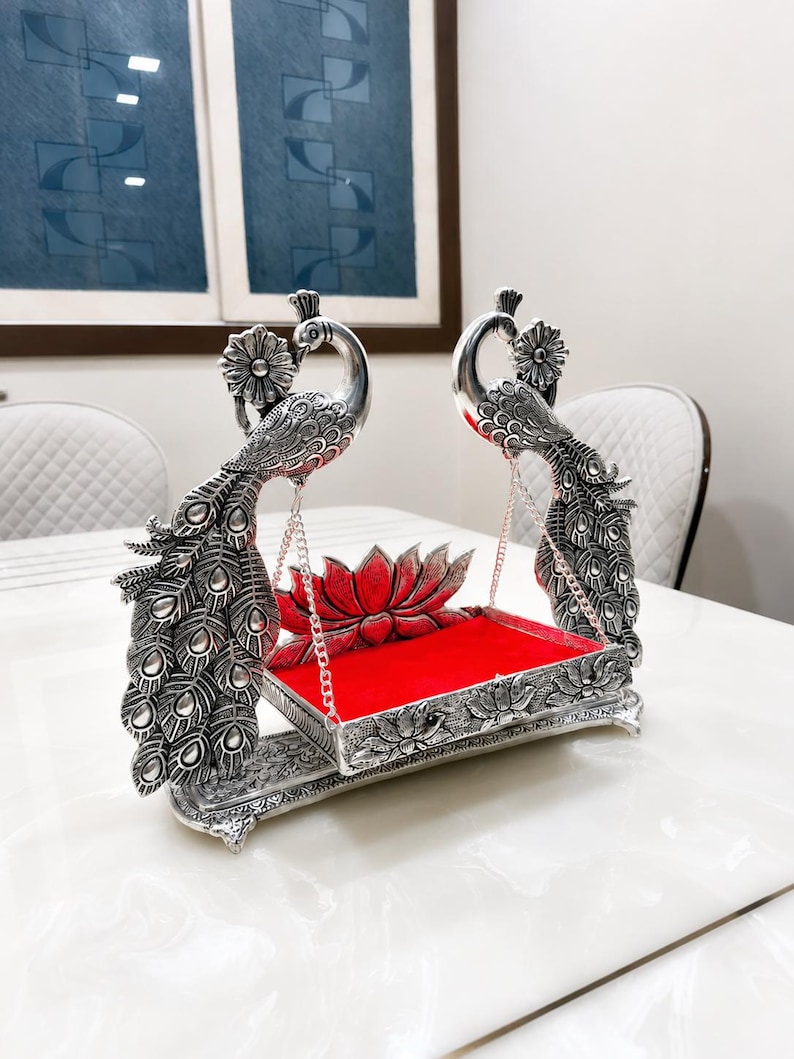 May include: A silver-colored metal swing featuring two peacock figures with detailed feather patterns on the sides. The swing has a rectangular base with a bright red velvet-like insert and a lotus flower motif at the back, suspended by chains.