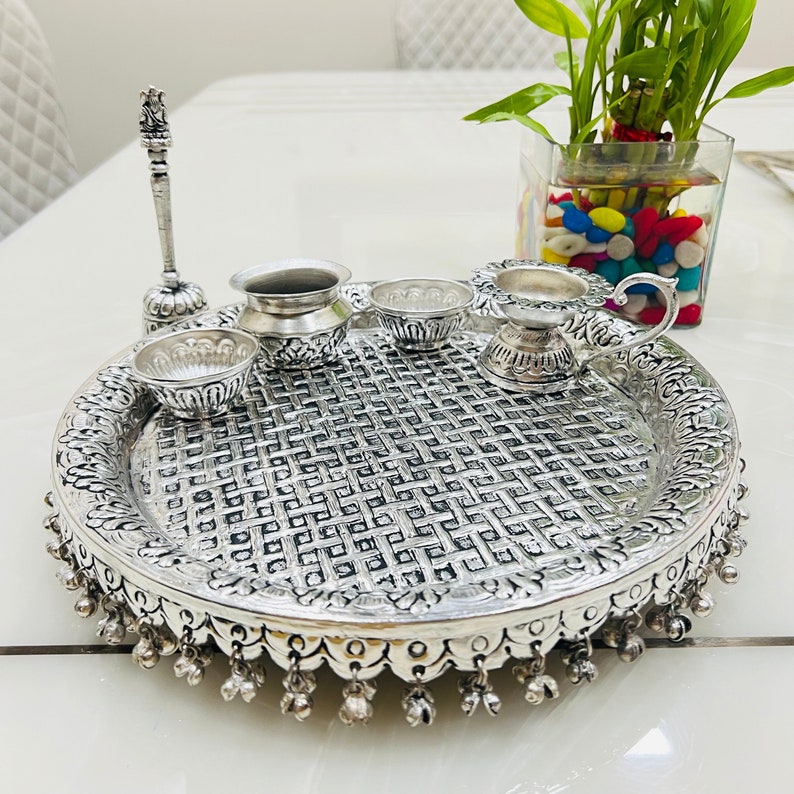 May include: A silver-plated puja thali with a decorative woven pattern, featuring a small silver pitcher, a diya, and three small bowls. The thali has a decorative border with small bells.