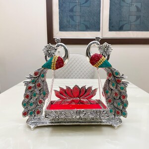 May include: A silver-toned decorative swing featuring two peacock figures with red and teal accents on their heads and tails. The central platform is rectangular with a red velvet base, positioned in front of a sculpted silver lotus flower element.