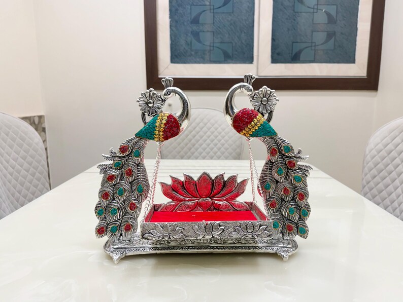 May include: A silver-toned decorative swing featuring two peacock figures with red and teal accents on their heads and tails. The central platform is rectangular with a red velvet base, positioned in front of a sculpted silver lotus flower element.