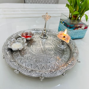 May include: A silver tray with intricate floral designs, featuring a small silver bell, a bowl with red liquid, a bowl with white rice, and a lit oil lamp.