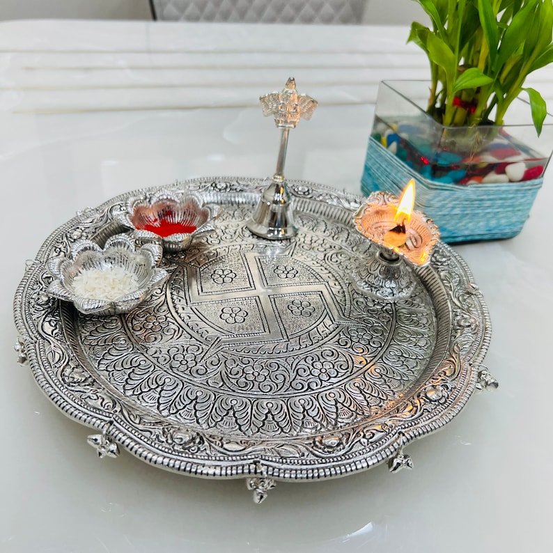May include: A silver tray with intricate floral designs, featuring a small silver bell, a bowl with red liquid, a bowl with white rice, and a lit oil lamp.