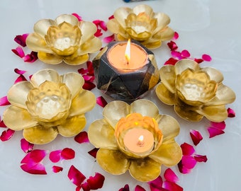 May include: Five gold metal lotus flower candle holders with a black stone candle holder in the center. The candle holders are arranged in a circle on a white surface with pink flower petals scattered around them.