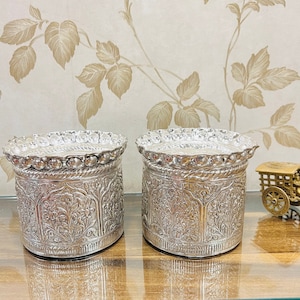 May include: Two silver decorative containers with intricate floral designs. The containers have a round top and a cylindrical body. The containers are on a wooden surface.