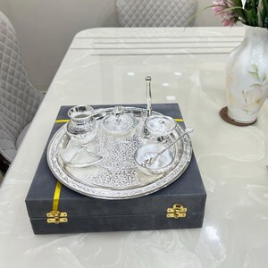 May include: A silver serving tray with a decorative design, containing a small silver pitcher, two small bowls with lids, a small bowl, and a spoon. The tray is in a black box with a yellow ribbon.