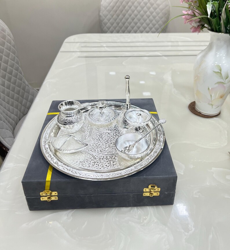 May include: A silver serving tray with a decorative design, containing a small silver pitcher, two small bowls with lids, a small bowl, and a spoon. The tray is in a black box with a yellow ribbon.