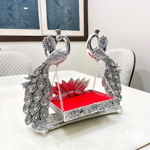 May include: A silver-toned metal swing featuring two peacock figures with detailed feather patterns. The swing holds a rectangular platform with a bright red velvet lining and a decorative lotus flower accent at the back, suspended by thin silver chains.