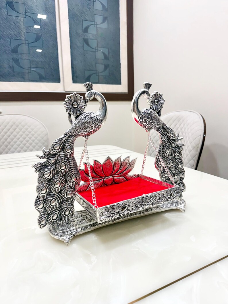 May include: A silver-toned metal swing featuring two peacock figures with detailed feather patterns. The swing holds a rectangular platform with a bright red velvet lining and a decorative lotus flower accent at the back, suspended by thin silver chains.