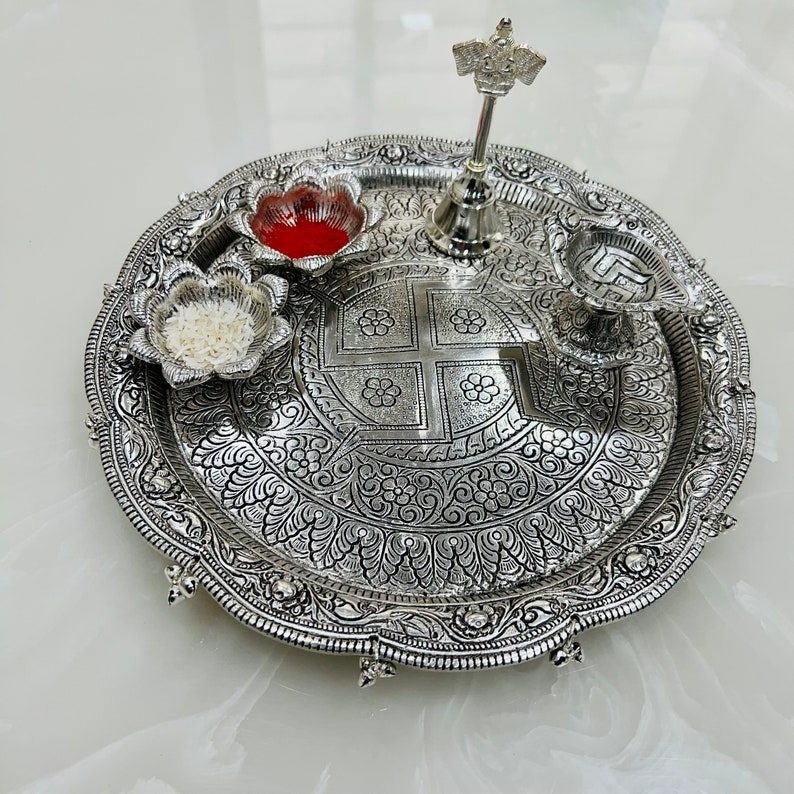 May include: A silver-plated puja thali with intricate floral designs. It features four small bowls, a bell, and a decorative symbol in the center. The thali is perfect for religious ceremonies and rituals.
