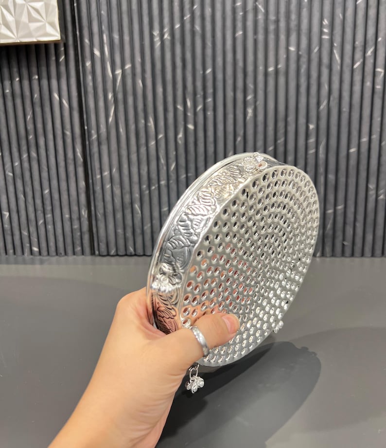 May include: Silver round tray with a decorative floral design and a perforated bottom. The tray has three small bells hanging from the bottom.
