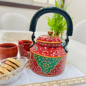 May include: A red metal teapot with a black handle and intricate hand-painted floral designs in gold, green, and white. The teapot is sitting on a white table with a placemat.