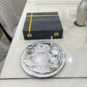May include: A silver serving tray with an intricate design, featuring a small silver pitcher, a bowl, a lidded container, a small cup, and a spoon. The tray is on a white table with a gray box with a yellow ribbon.
