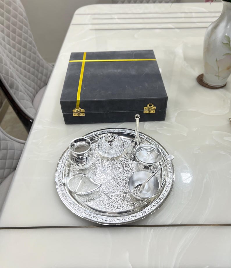 May include: A silver serving tray with an intricate design, featuring a small silver pitcher, a bowl, a lidded container, a small cup, and a spoon. The tray is on a white table with a gray box with a yellow ribbon.