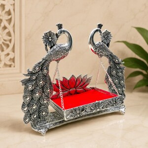 May include: A silver-toned metal swing featuring two peacock figures with detailed feather patterns on either side. The swing platform is rectangular with a bright red velvet base and a red lotus flower ornament at the back, suspended by silver chains.