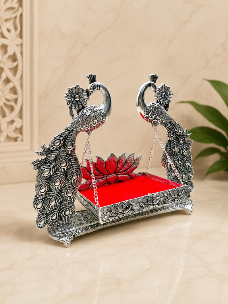 May include: A silver-toned metal swing featuring two peacock figures with detailed feather patterns on either side. The swing platform is rectangular with a bright red velvet base and a red lotus flower ornament at the back, suspended by silver chains.