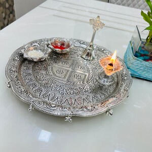 May include: A silver tray with intricate floral designs, featuring a small bowl of white powder, a small bowl of red liquid, a silver bell, and a silver oil lamp with a lit wick.