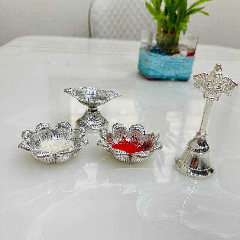 May include: A silver bell with a bird-like figure on top, two silver bowls with white and red powder, and a silver diya.