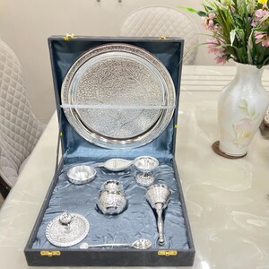 May include: A silver serving set in a black velvet box. The set includes a large round tray, a small bowl, a pitcher, a spoon, a small cup, and a small lidded dish. The tray and all the pieces have intricate floral designs.