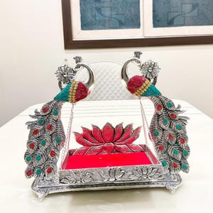 May include: A silver-toned metal swing featuring two peacock figures with red and turquoise inlaid accents. The central platform has a bright red velvet base and a lotus flower backrest, with the peacocks connected by silver chains.