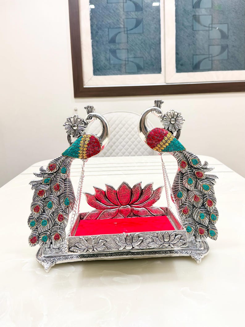 May include: A silver-toned metal swing featuring two peacock figures with red and turquoise inlaid accents. The central platform has a bright red velvet base and a lotus flower backrest, with the peacocks connected by silver chains.