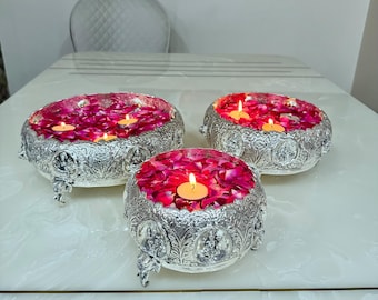 Puede incluir: Tres cuencos redondos de metal plateado con intrincados detalles en relieve y patas pequeñas. Cada cuenco está lleno de pétalos de rosa de color rosa vibrante y contiene pequeñas velas de té encendidas, creando un brillo cálido en los recipientes.