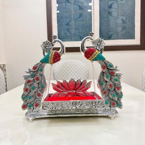 May include: A silver-toned decorative swing featuring two peacocks with red and turquoise accents on their wings and tails. The central platform is rectangular with a red velvet base and a lotus flower motif, suspended by chains between the peacocks.