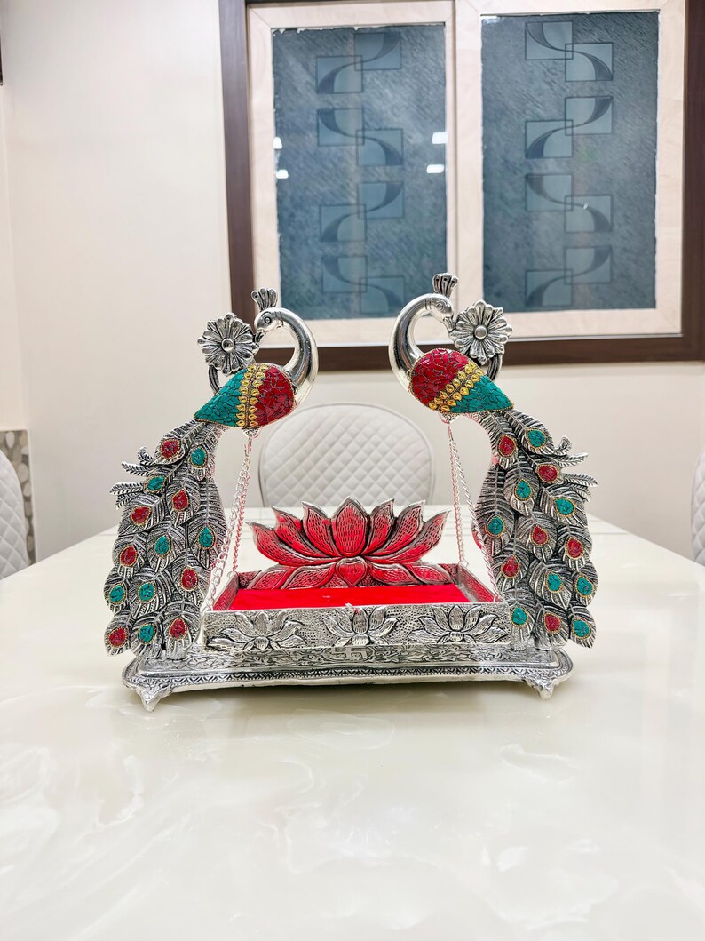 May include: A silver-toned decorative swing featuring two peacocks with red and turquoise accents on their wings and tails. The central platform is rectangular with a red velvet base and a lotus flower motif, suspended by chains between the peacocks.