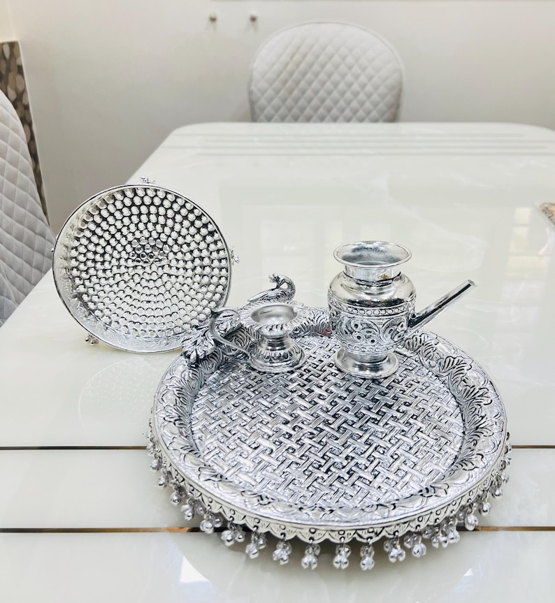 May include: A silver tray with intricate designs, featuring a small silver pitcher, a diya, and a peacock design. The tray has a beaded border.