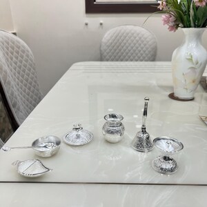 May include: A set of six silver-plated ceremonial items, including a small pitcher, a bowl, a lidded dish, a spoon, a bell, and a small cup. The items are arranged on a white table.