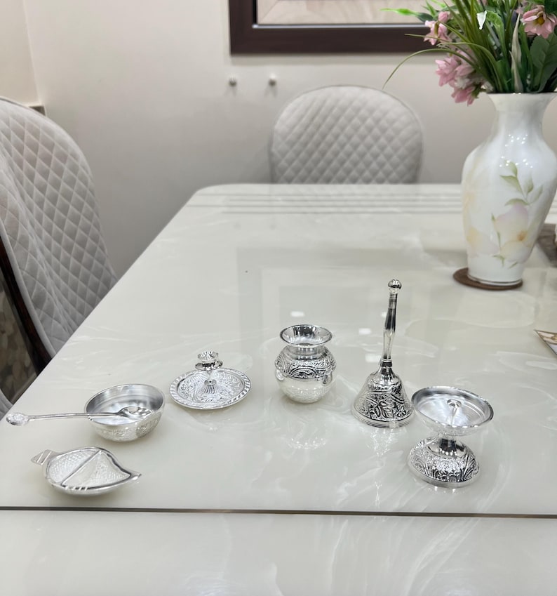 May include: A set of six silver-plated ceremonial items, including a small pitcher, a bowl, a lidded dish, a spoon, a bell, and a small cup. The items are arranged on a white table.