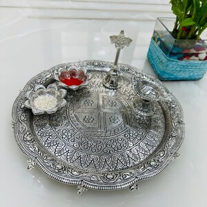 May include: A silver-colored metal plate with intricate floral designs. The plate has four small bowls, one with red powder, one with white grains, and two empty. A silver bell with a decorative top sits in the center of the plate.