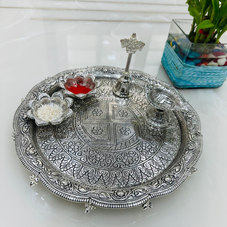 May include: A silver-colored metal plate with intricate floral designs. The plate has four small bowls, one with red powder, one with white grains, and two empty. A silver bell with a decorative top sits in the center of the plate.