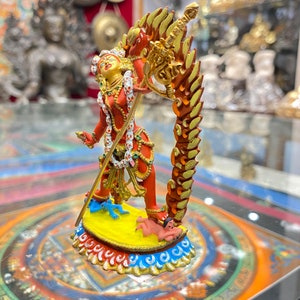 May include: A red and gold statue of a deity with a sword and a third eye. The deity is standing on a yellow base with blue and white trim. The statue is surrounded by a colorful mandala.