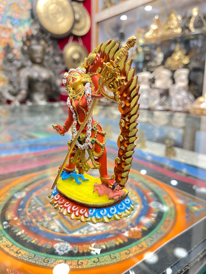 May include: A red and gold statue of a deity with a sword and a third eye. The deity is standing on a yellow base with blue and white trim. The statue is surrounded by a colorful mandala.