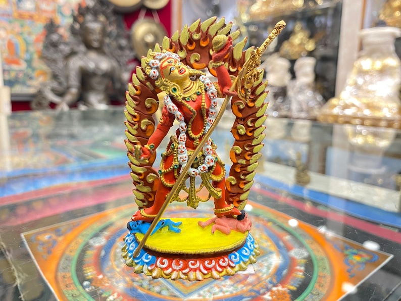 May include: A red and gold statue of a deity with a fierce expression, holding a spear and standing on two figures. The statue is surrounded by a halo of flames.